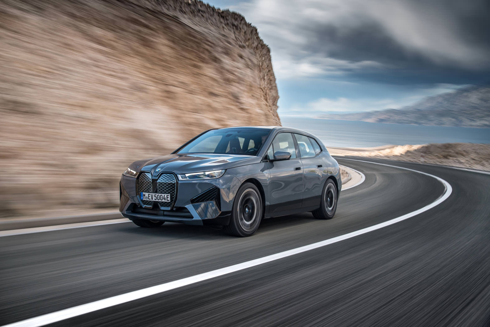 Shot of the BMW iX driving along a road, background is blurred and the sea is visible in the distance.