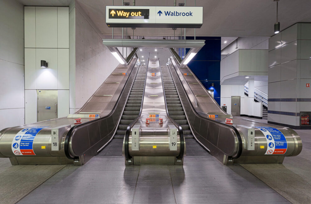 Crop from a 360 showing the view looking up 2 escalators at a tube station. There is a large sign showing'Way Out' and'Walbrook' with arrows pointing up the escalator visible above.