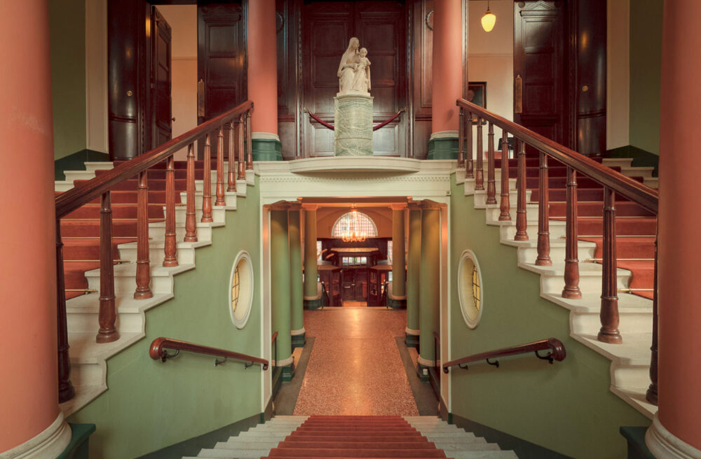 View inside Archbishop's House from our Westminster Cathedral virtual tour. This image is a still showing a view looking down a grand, staircase into a loby with large columns. At the sides are two further staircases leading to a gallery above with a statue of the virgin Mary and child.