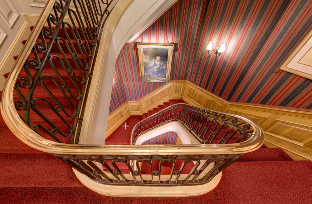 Stairwell image taken from the apartment virtual tours for 47 Park Street. This shows a grand, curved staircase, carpeted in red and with red-papered and wood-panelled walls, hung with gilt-framed artwork.